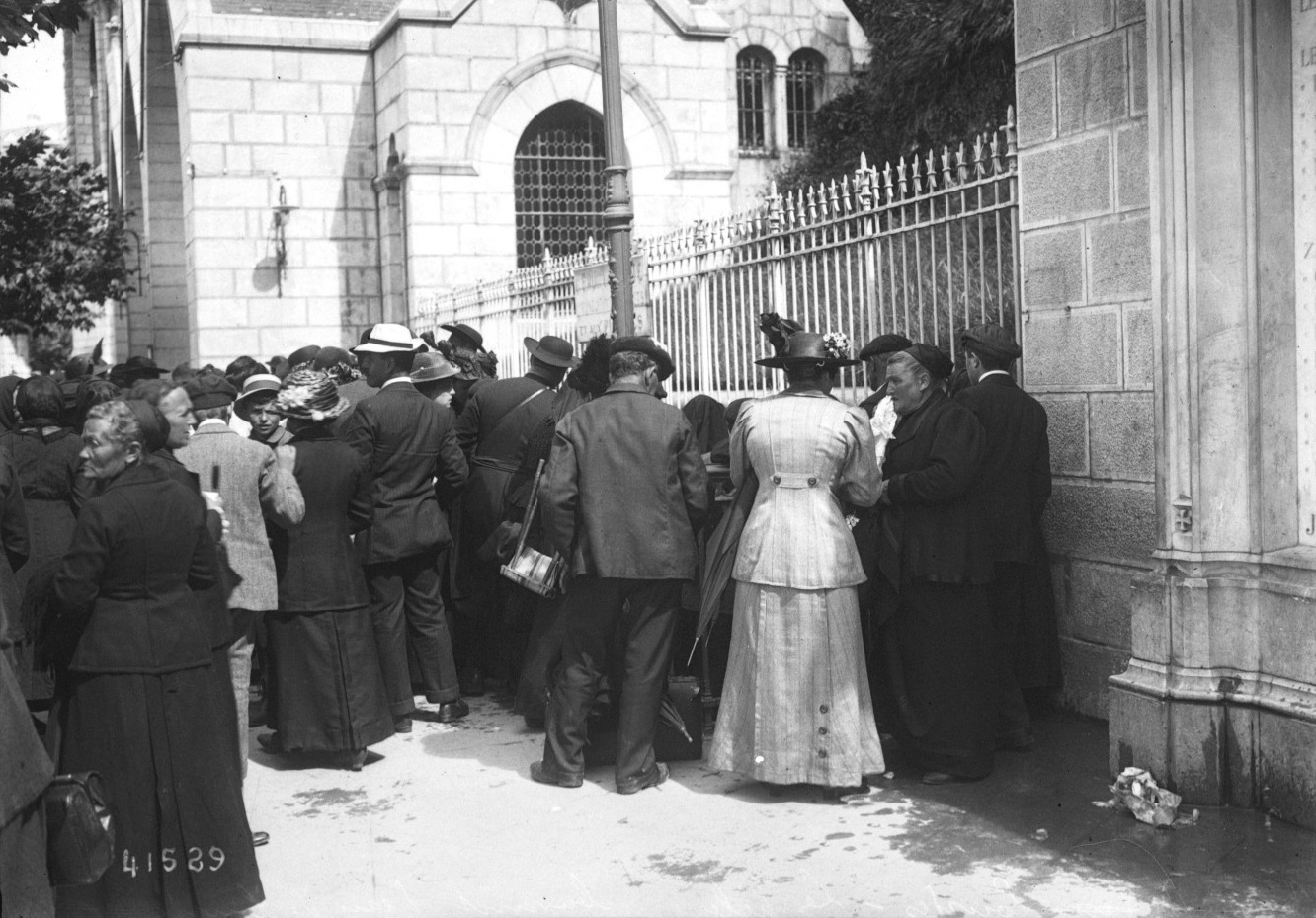 Fotografía de 1914 de una multitud de peregrinos, hombres con sombrero y mujeres con vestidos largos, apiñados junto a una verja delante de un edificio de piedra