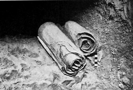 Fotografía en blanco y negro de dos rollos enrollados que yacen sobre el suelo de piedra de una cueva, junto a una escala métrica