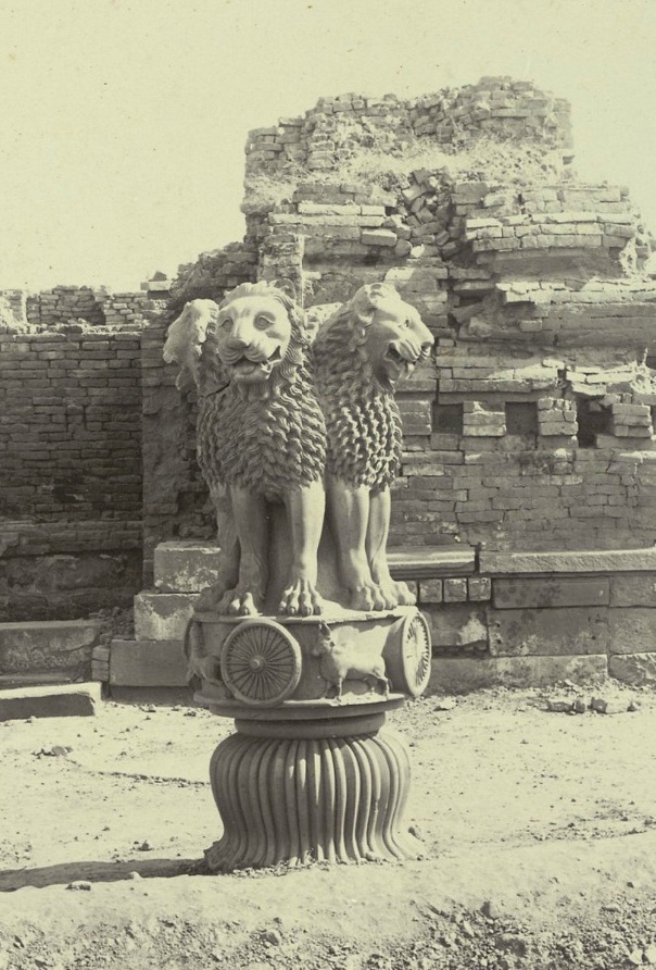 Fotografía antigua de un capitel de piedra con cuatro leones sentados espalda con espalda sobre una base circular, delante de unas ruinas de ladrillo