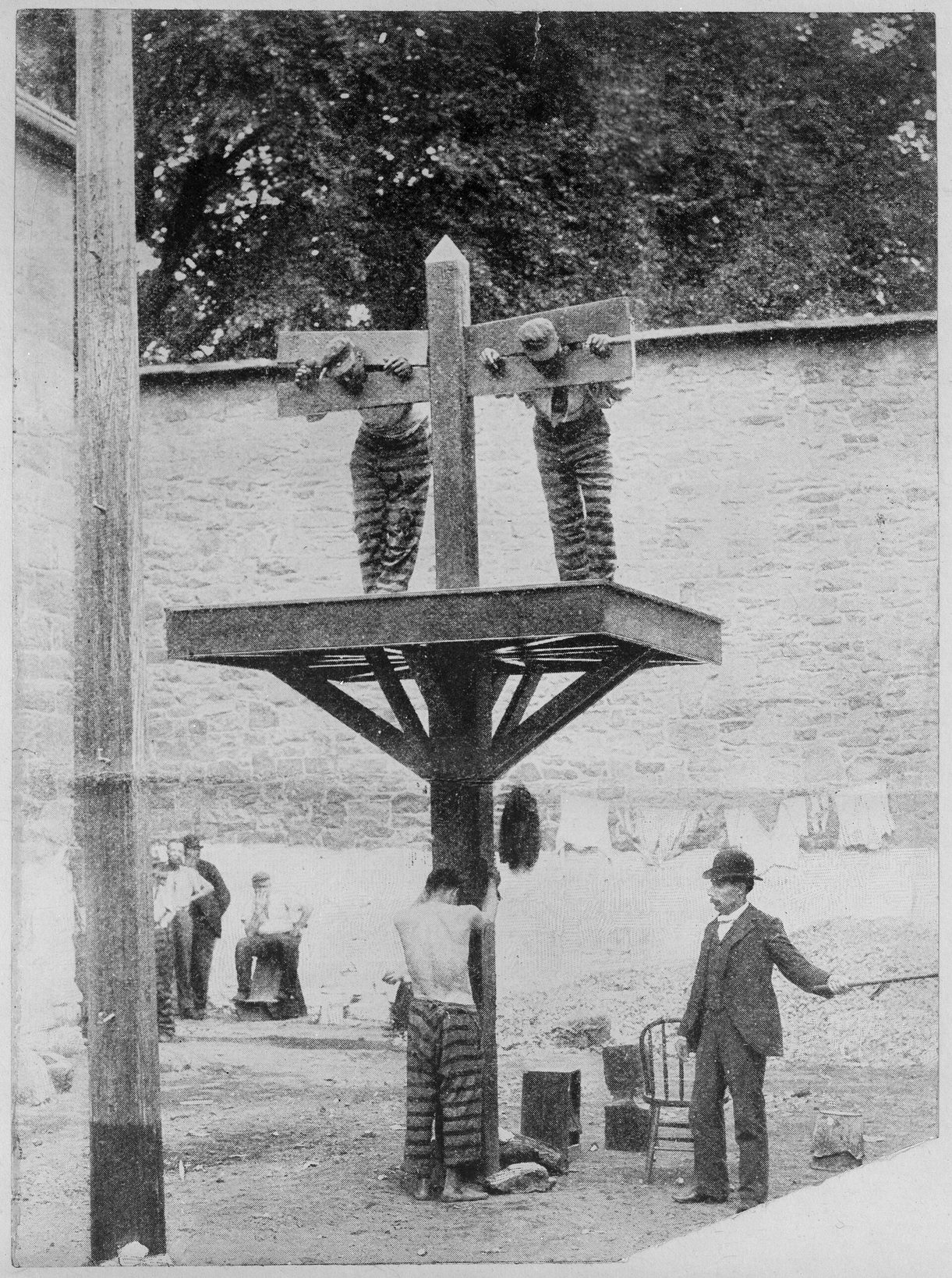 Fotografía antigua en blanco y negro de dos presos con uniforme de rayas inmovilizados en una picota de madera sobre un poste, con otro atado abajo y varios hombres alrededor en un patio