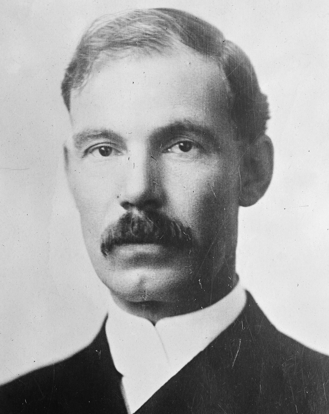 Retrato en blanco y negro de un hombre de mediana edad con bigote poblado, pelo peinado con raya y cuello alto blanco