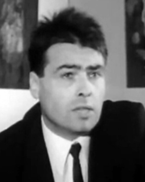 Fotograma en blanco y negro de un hombre joven de pelo oscuro, con chaqueta, camisa blanca y corbata, que mira hacia un lado mientras habla