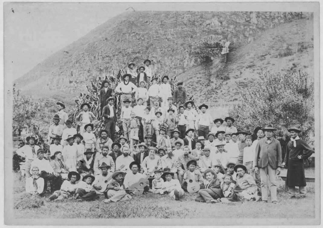 Fotografía antigua en blanco y negro de un numeroso grupo de personas, adultos y niños, sentadas y de pie al aire libre al pie de una ladera