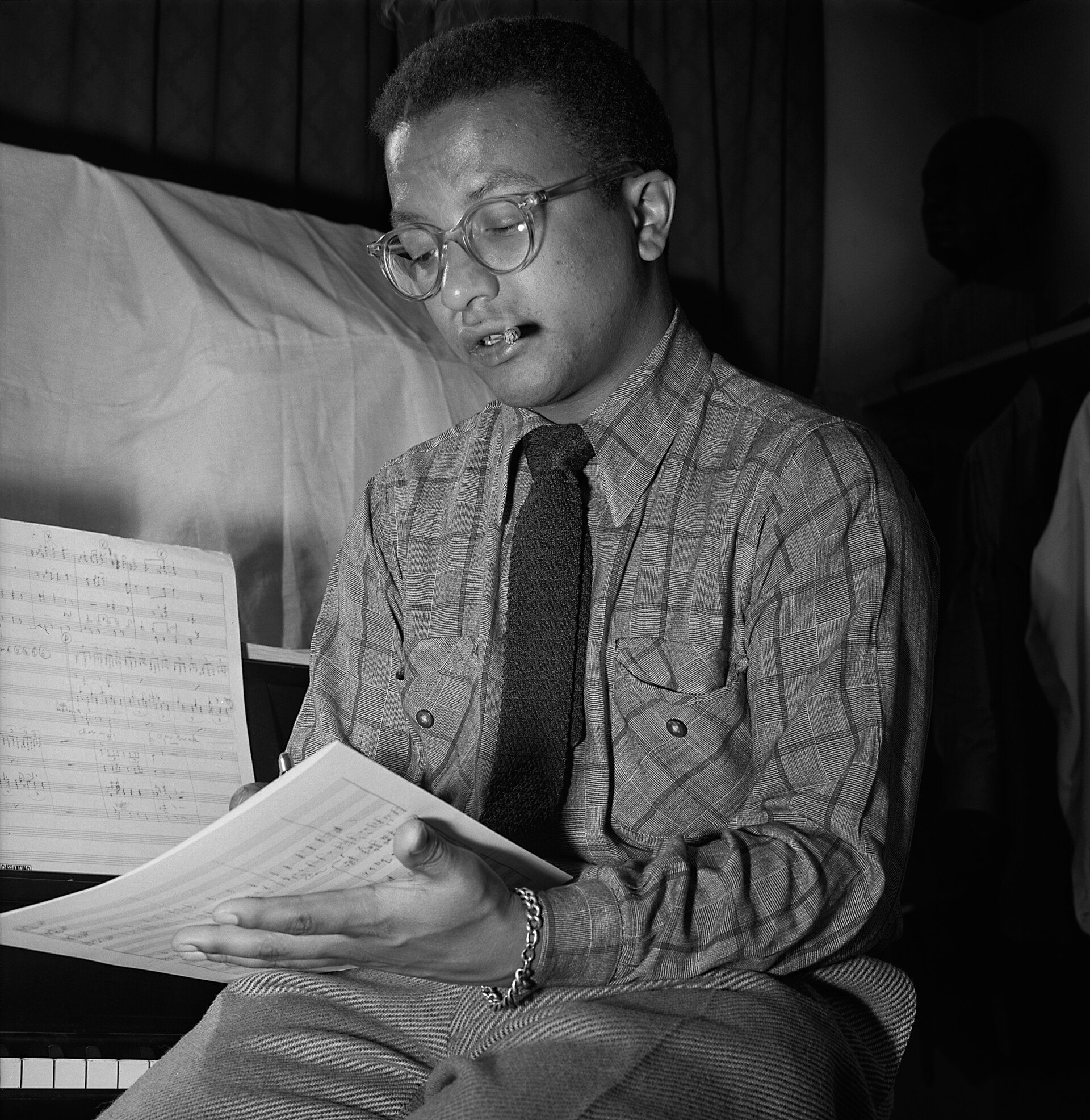 Fotografía en blanco y negro de un hombre joven con gafas, camisa de cuadros y corbata, sentado ante un piano mientras lee una partitura