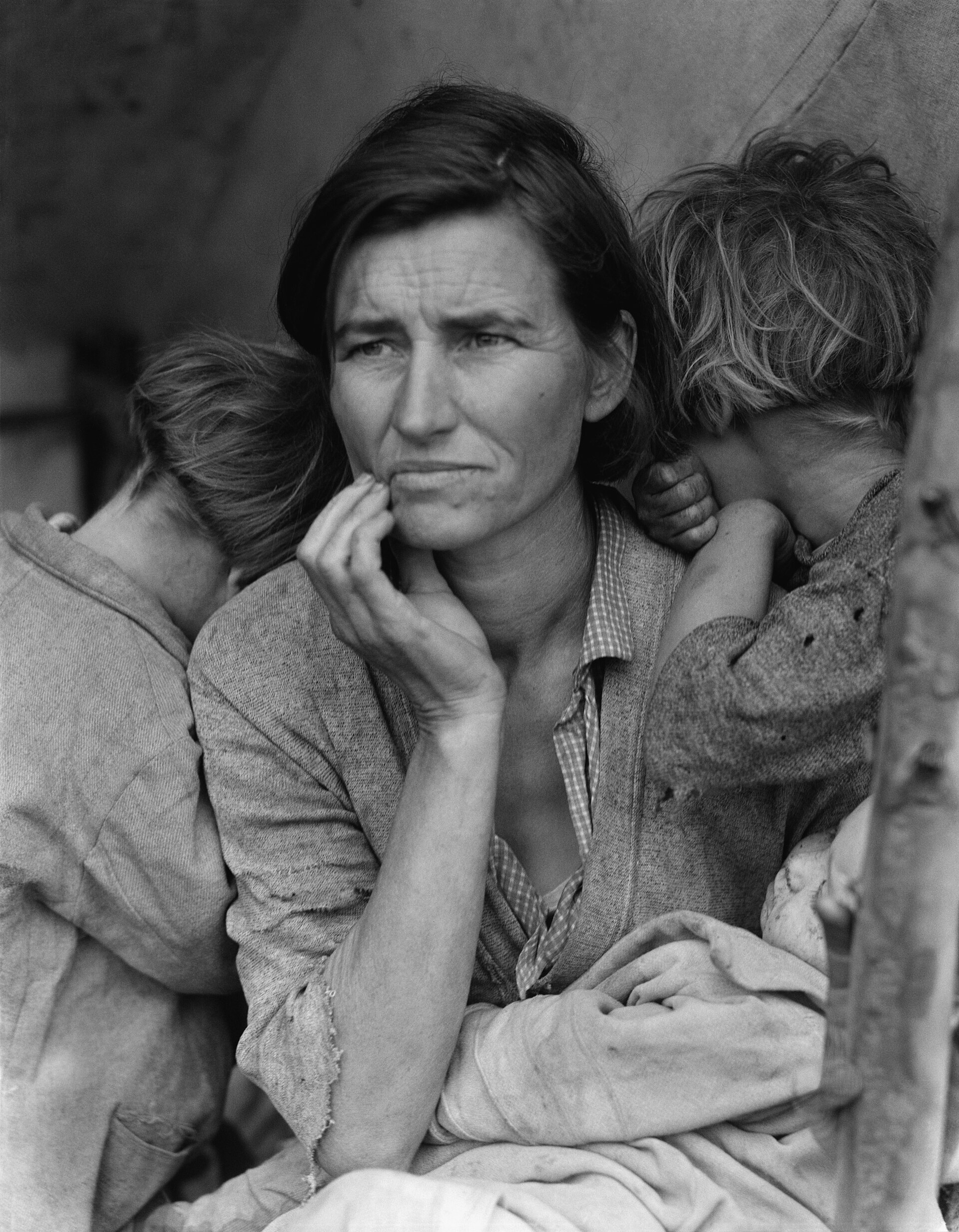 Fotografía en blanco y negro de una mujer de rostro preocupado que se lleva la mano a la barbilla, con dos niños apoyados en sus hombros de espaldas a la cámara y un bebé en brazos