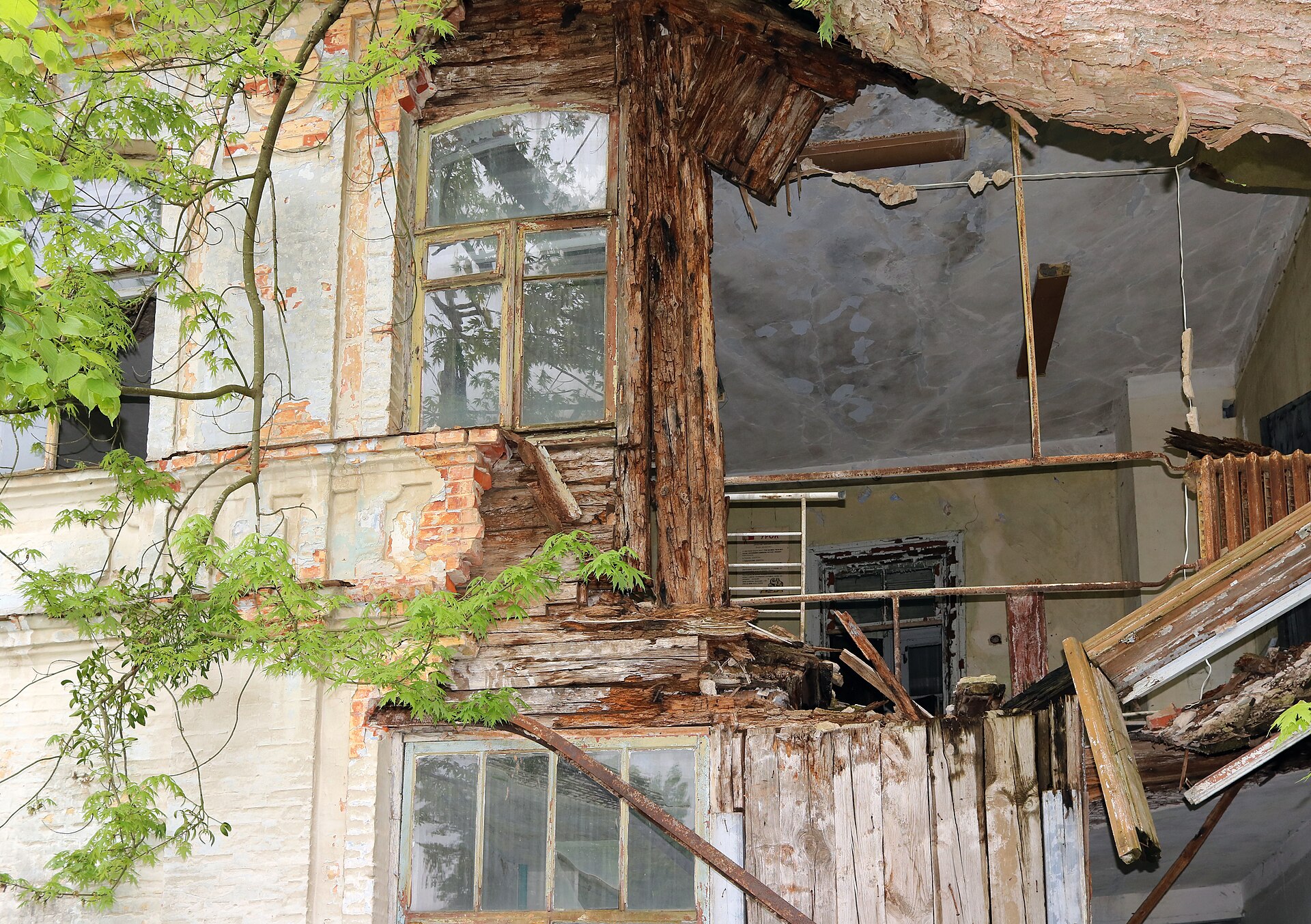Fotografía en color de un edificio de madera y ladrillo en ruinas, con el tejado hundido y la vegetación creciendo entre los escombros