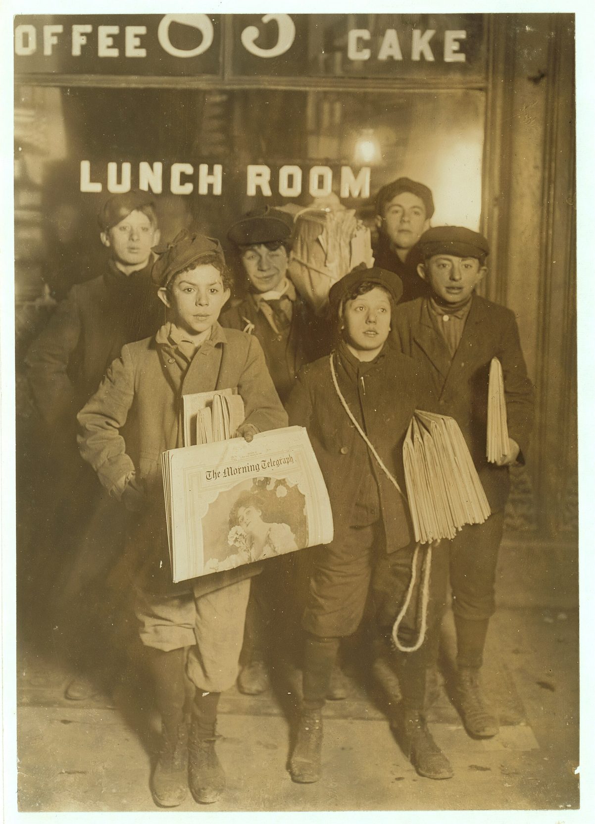 Fotografía de 1908 de un grupo de chicos con fajos de periódicos bajo el brazo posando de noche ante la puerta iluminada de un local