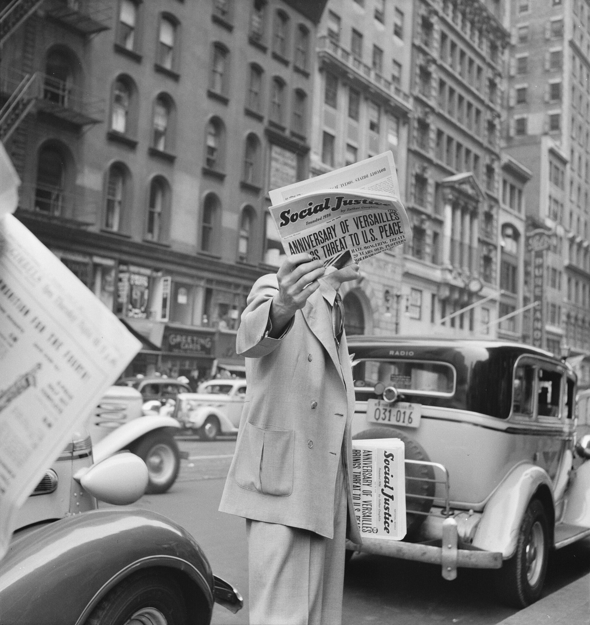 Fotografía de 1939 de un hombre en una calle sosteniendo un periódico abierto que le tapa la cara por completo, junto a coches aparcados