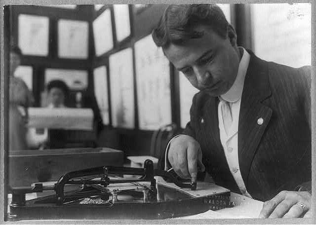 Fotografía de hacia 1908 de un empleado inclinado sobre un pantógrafo perforador, marcando una tarjeta sobre una mesa con documentos colgados en la pared