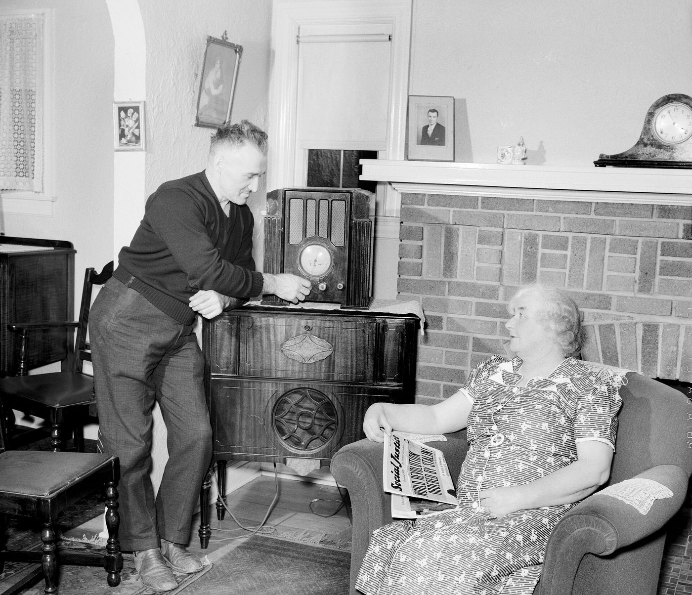 Fotografía de 1939 de un hombre ajustando un aparato de radio de madera en un salón mientras una mujer sentada en un sillón sostiene un periódico