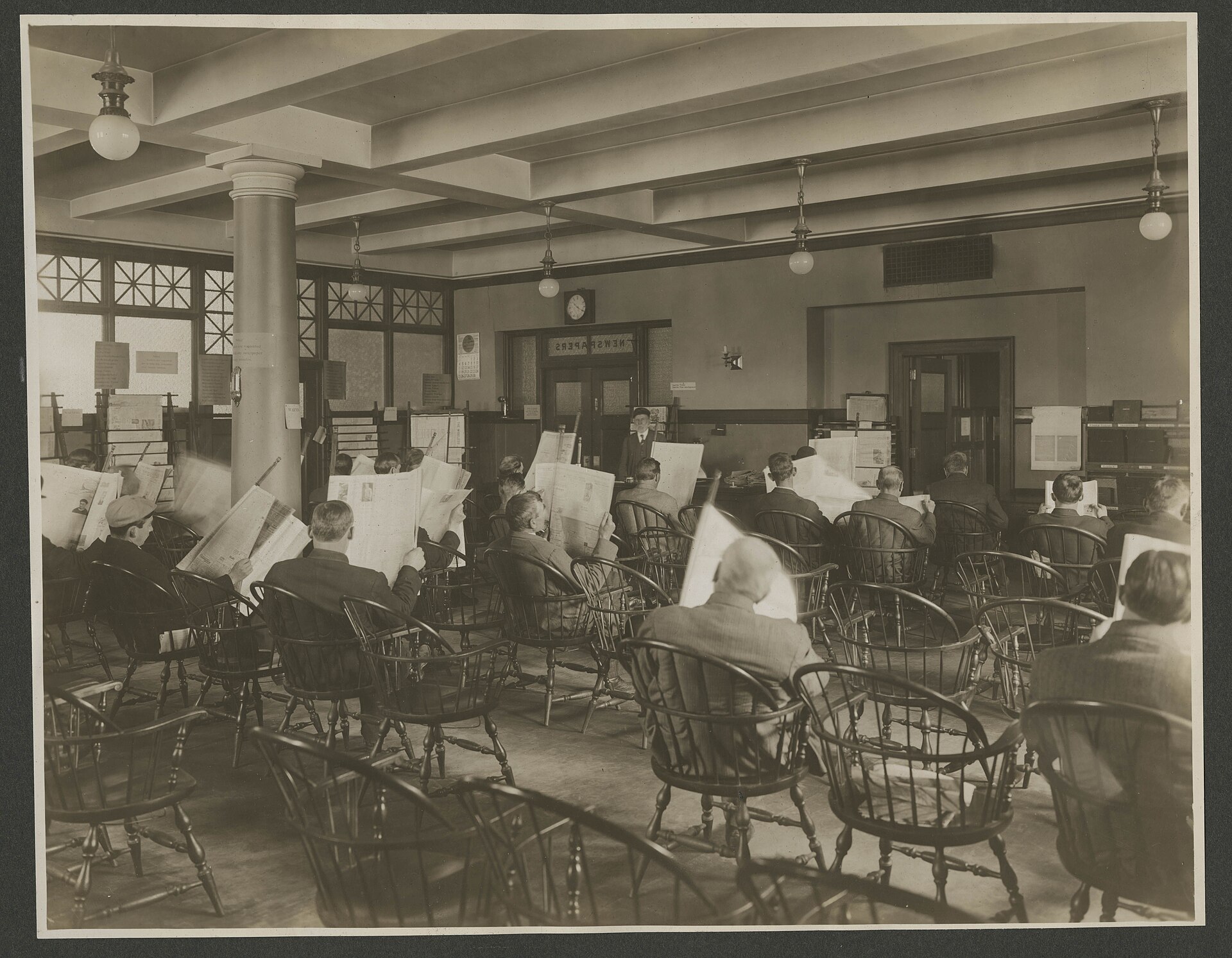 Fotografía de hacia 1910 de una gran sala de biblioteca con dos filas de hombres sentados en sillones, cada uno con un periódico abierto ante la cara
