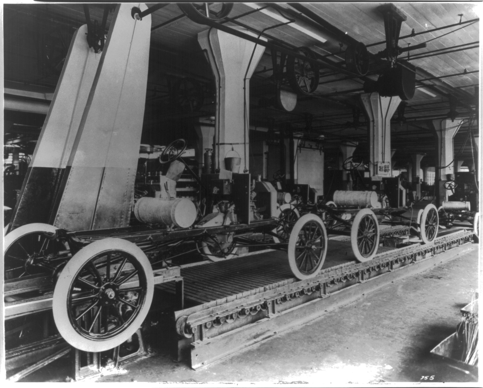 Fotografía de los años diez de una nave con chasis de automóvil alineados sobre una cinta y operarios trabajando a ambos lados