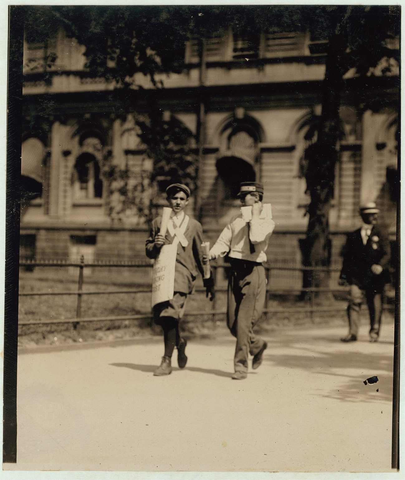 Fotografía de principios del siglo XX de dos chicos uniformados de mensajero caminando por una acera ante un edificio público