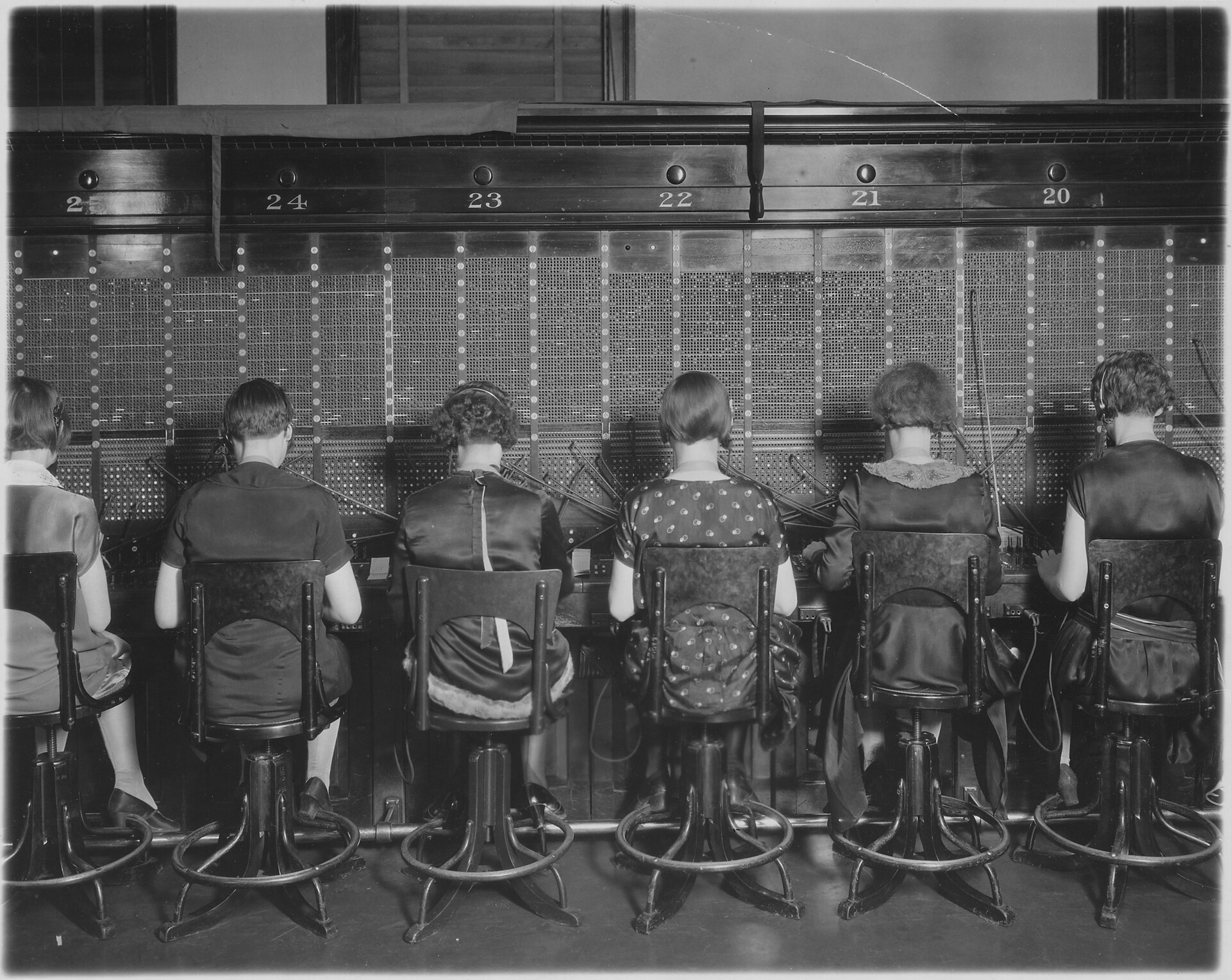 Fotografía de principios del siglo XX de una larga hilera de telefonistas de espaldas ante un cuadro de conmutación, sentadas en taburetes idénticos