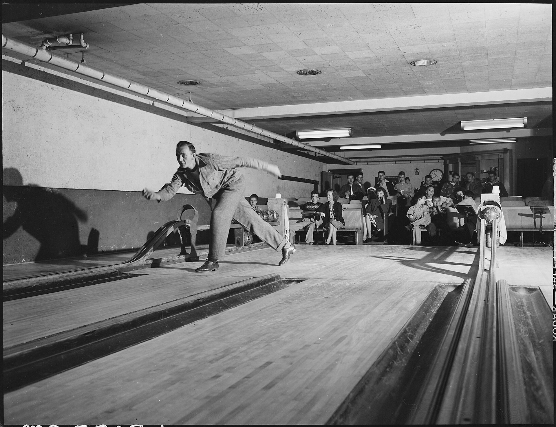 Fotografía de los años cuarenta de un joven lanzando una bola en una bolera mientras varios espectadores sentados al fondo observan