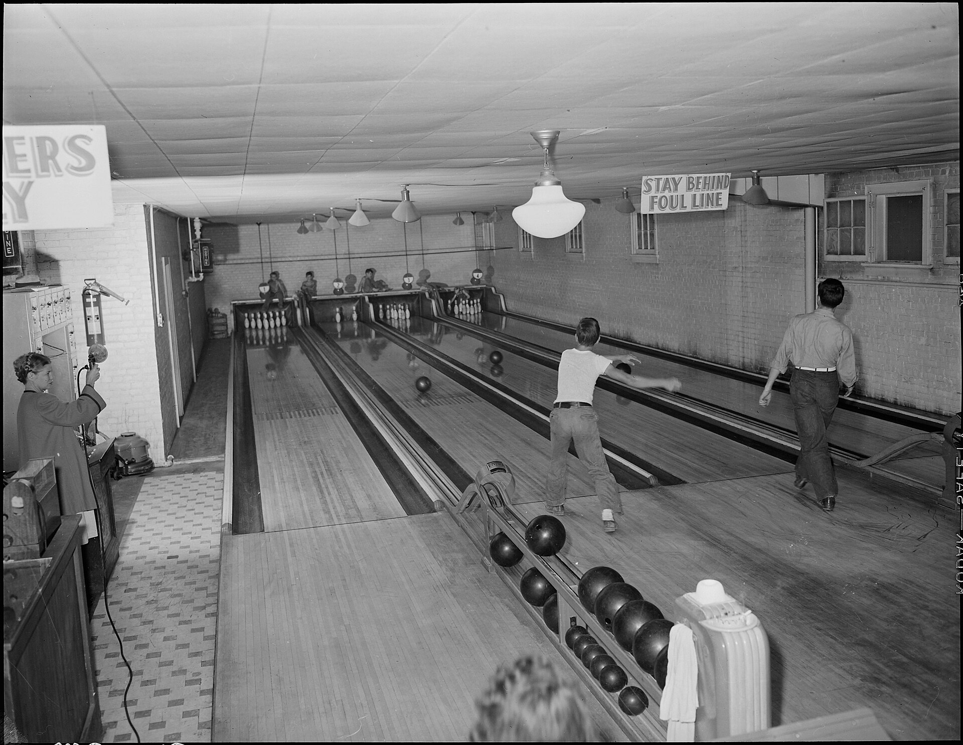 Fotografía de los años cuarenta de una bolera de dos pistas en un local comunitario, con un jugador lanzando y otro esperando al fondo