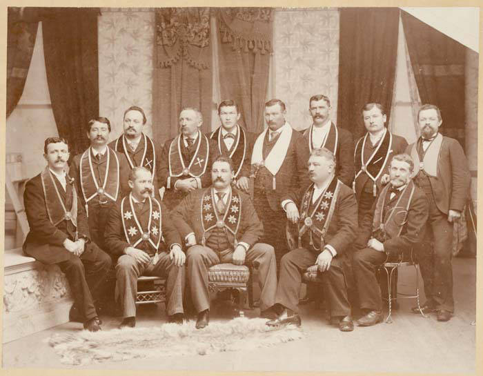 Retrato de estudio de hacia 1904 con nueve hombres de traje oscuro posando en dos filas, todos con bandas y medallas de una sociedad fraternal