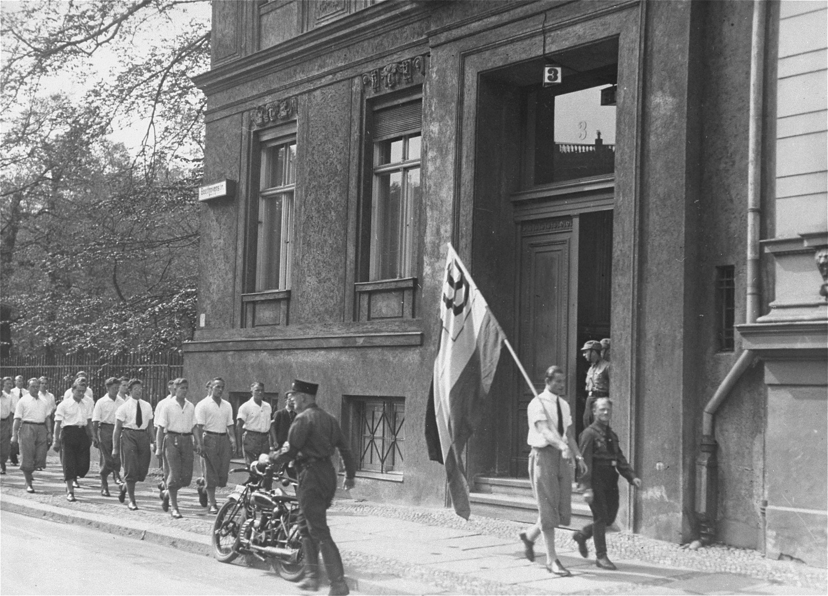 Fotografía de 1933 de una columna de jóvenes uniformados marchando ante un edificio con una bandera desplegada