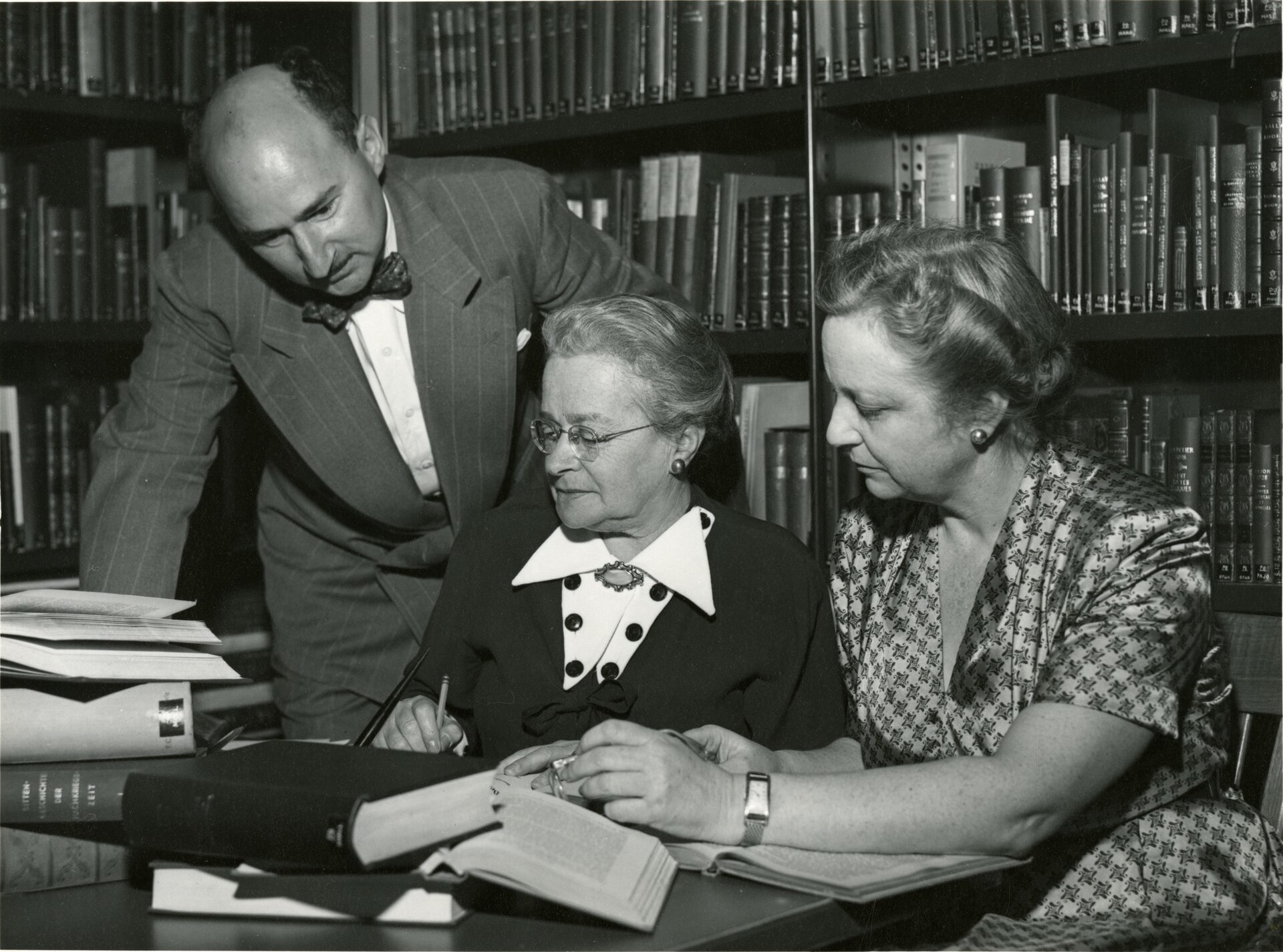 Fotografía en blanco y negro de tres personas trabajando con papeles y fichas alrededor de una mesa, ante estanterías llenas de libros