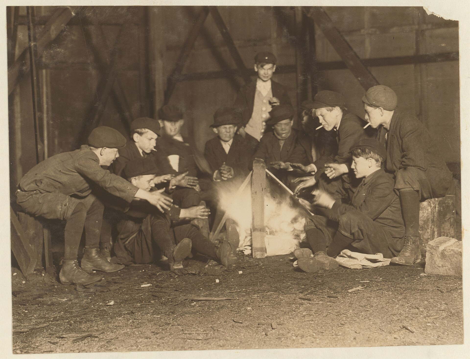 Fotografía nocturna de 1910 de una docena de chicos agrupados alrededor de una pequeña hoguera en un solar, detrás de una valla publicitaria