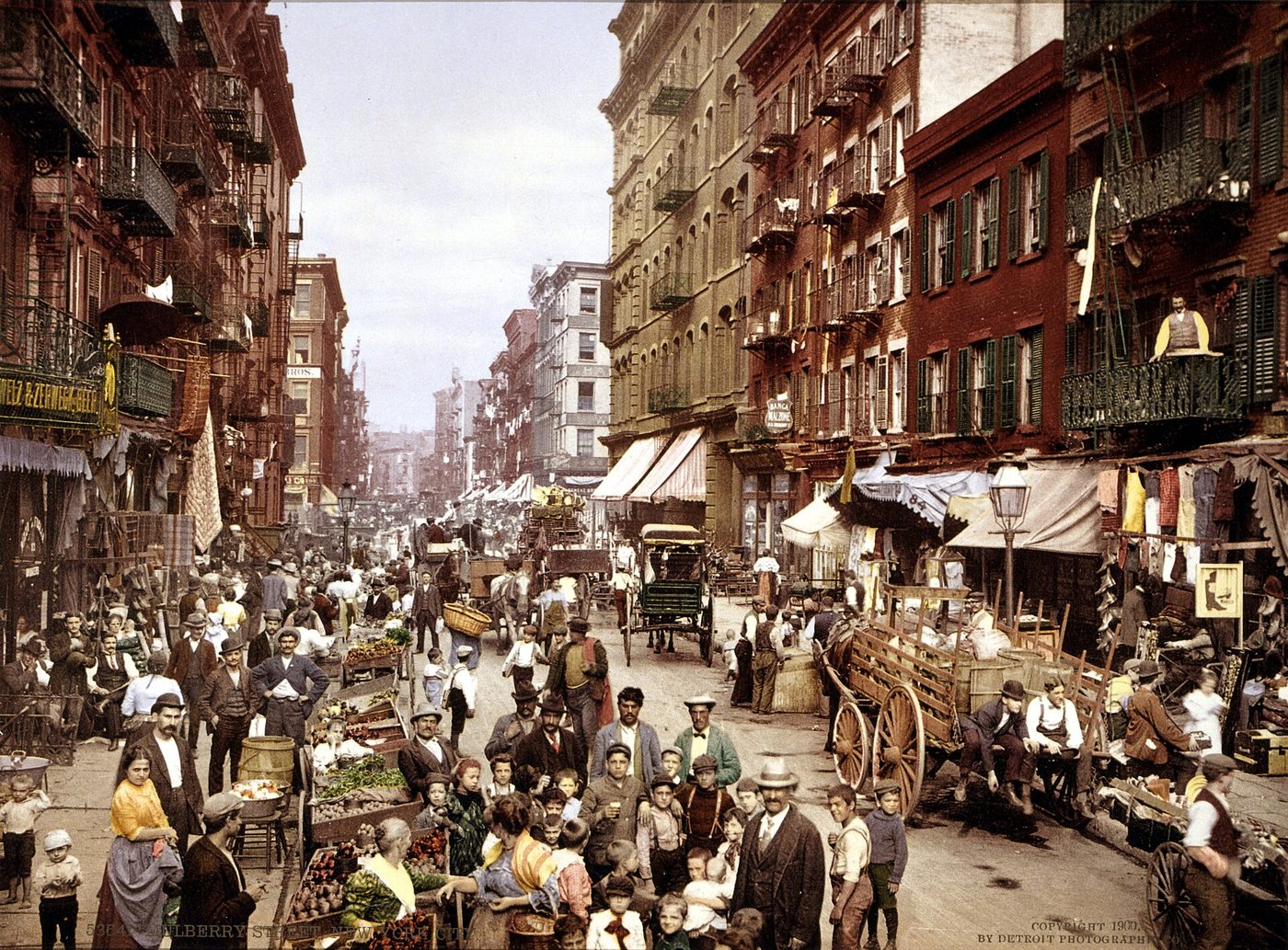 Fotografía en color de hacia 1900 de una calle estrecha de Nueva York abarrotada de puestos, carros y gente entre edificios de vecindad con escaleras de incendios