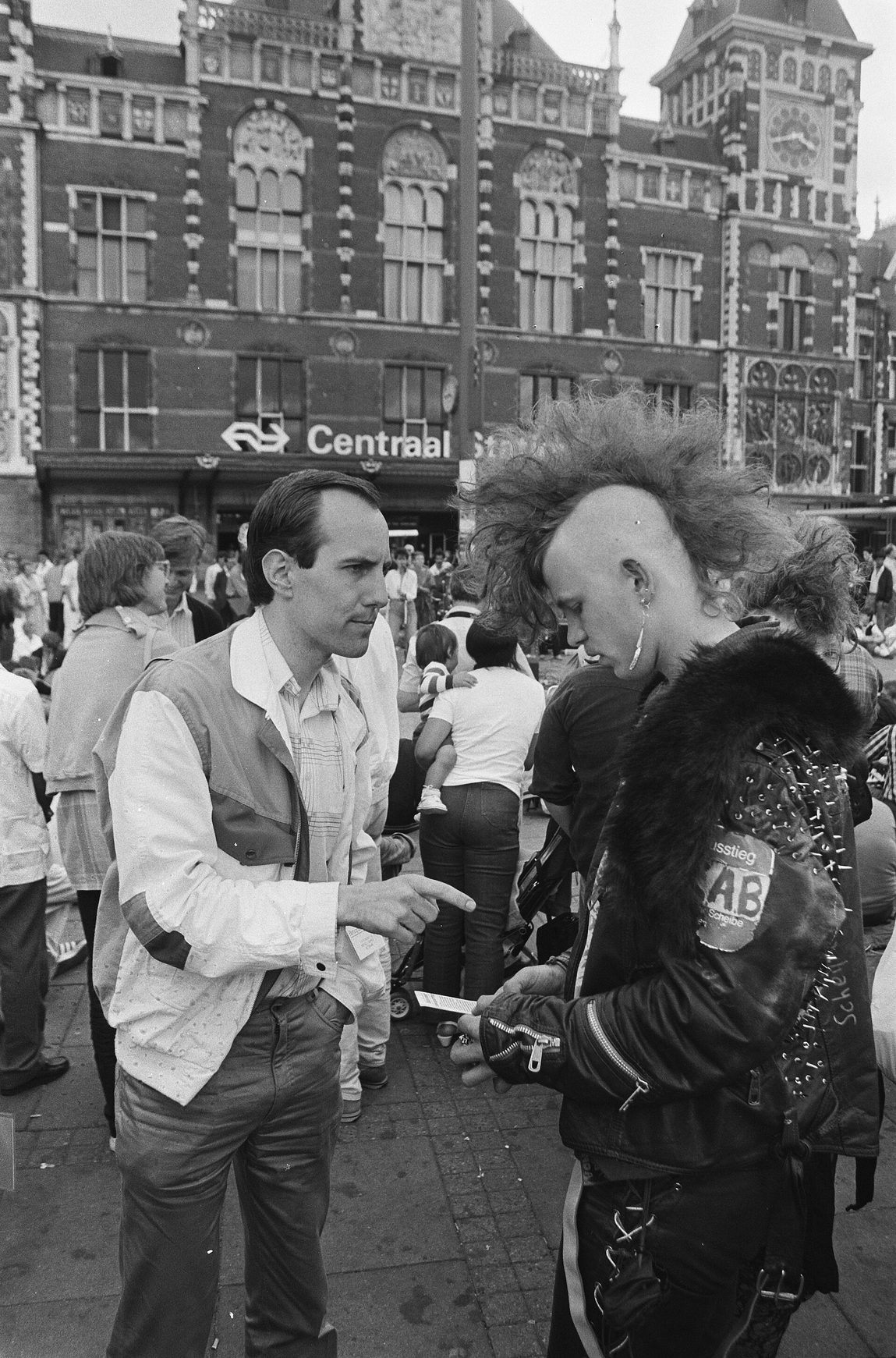 Fotografía de 1986 de un hombre con camisa blanca y folletos hablando con un joven de cresta y chaqueta de cuero en una calle concurrida