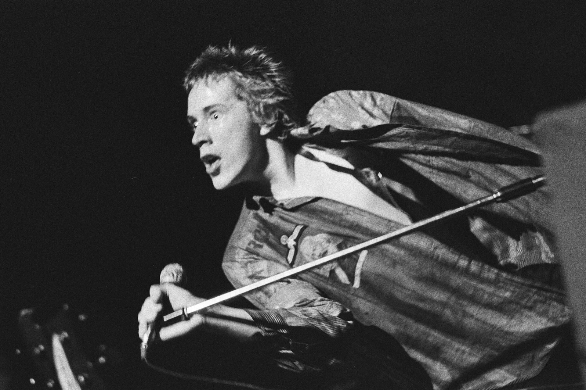 Fotografía en blanco y negro de enero de 1977 de un cantante joven inclinado sobre el micrófono, con chaqueta grande y gesto crispado, en un escenario oscuro