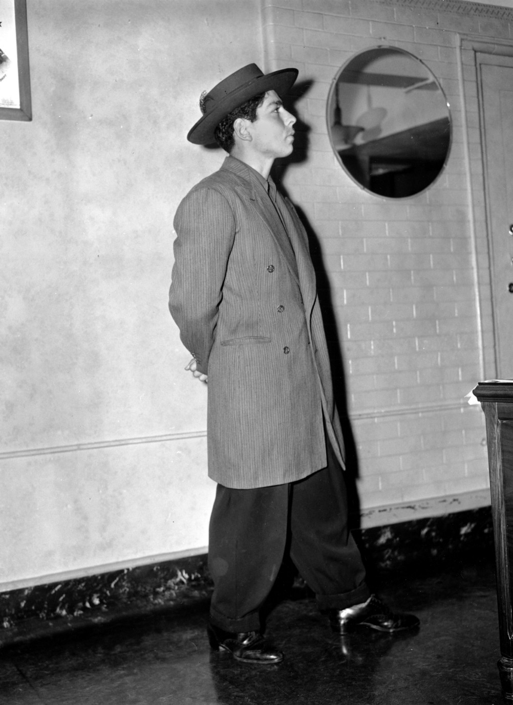 Fotografía de 1943 de un joven posando de perfil con traje de chaqueta larga y sombrero de ala ancha, dentro de una sala de azulejos