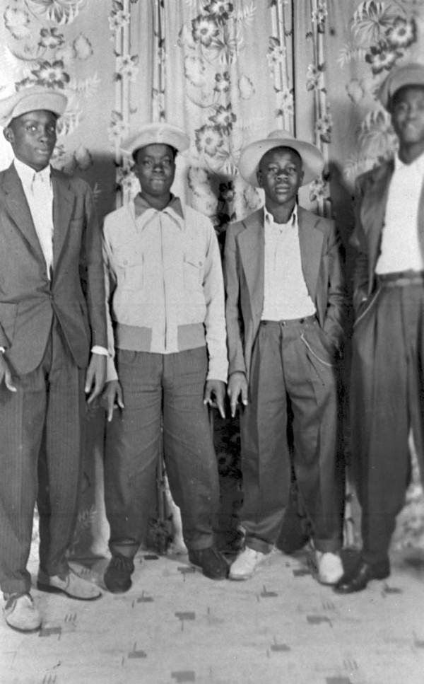Fotografía de 1940 de tres jóvenes posando sonrientes en la calle con trajes claros de chaqueta larga, pantalón ancho y cadenas colgando