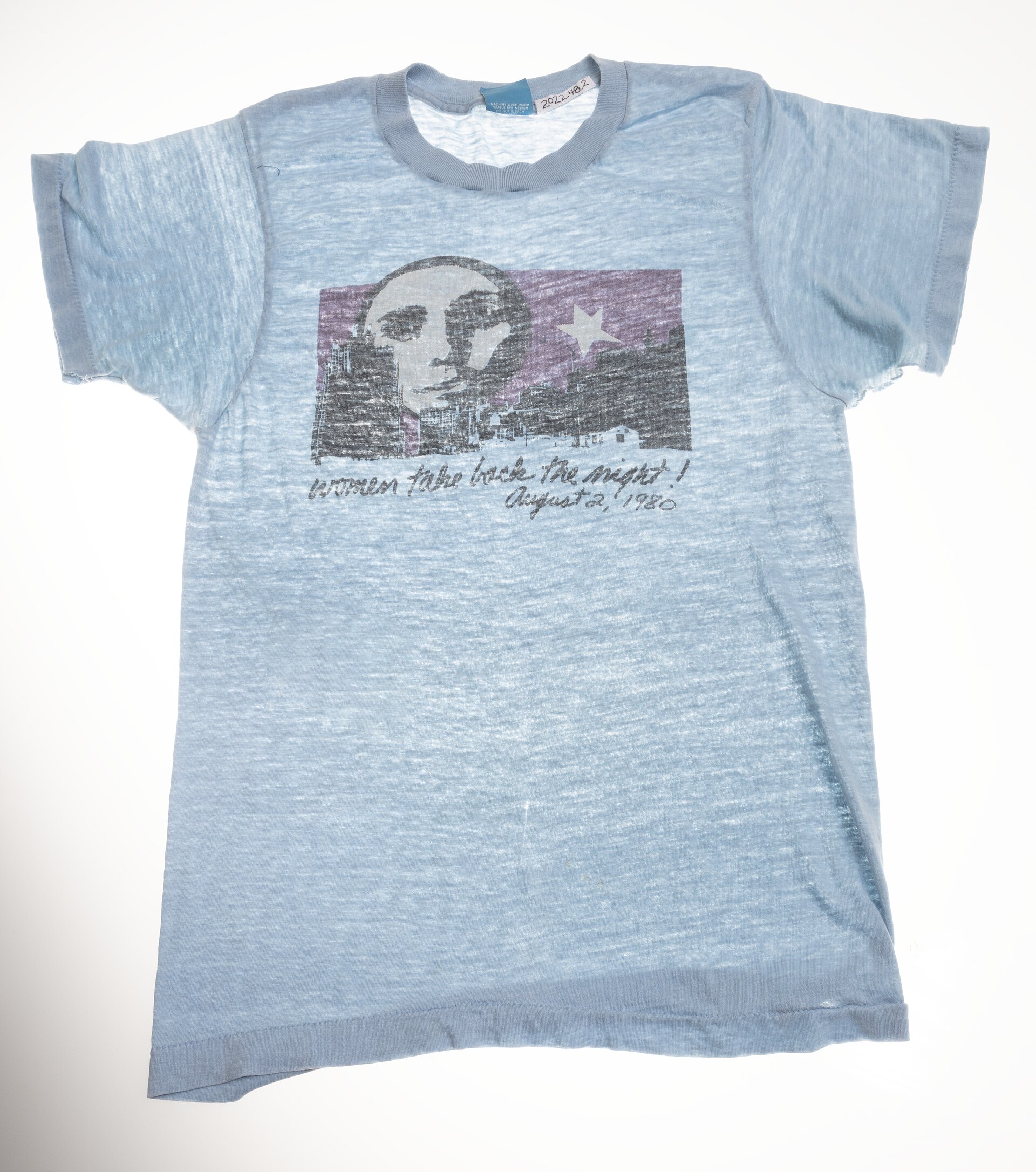 Camiseta azul claro desgastada con un dibujo del rostro de una mujer sobre fondo morado y el texto Women Take Back the Night