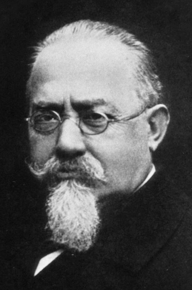 Retrato fotográfico en blanco y negro de un hombre mayor con gafas, bigote y barba blanca en punta, con traje oscuro