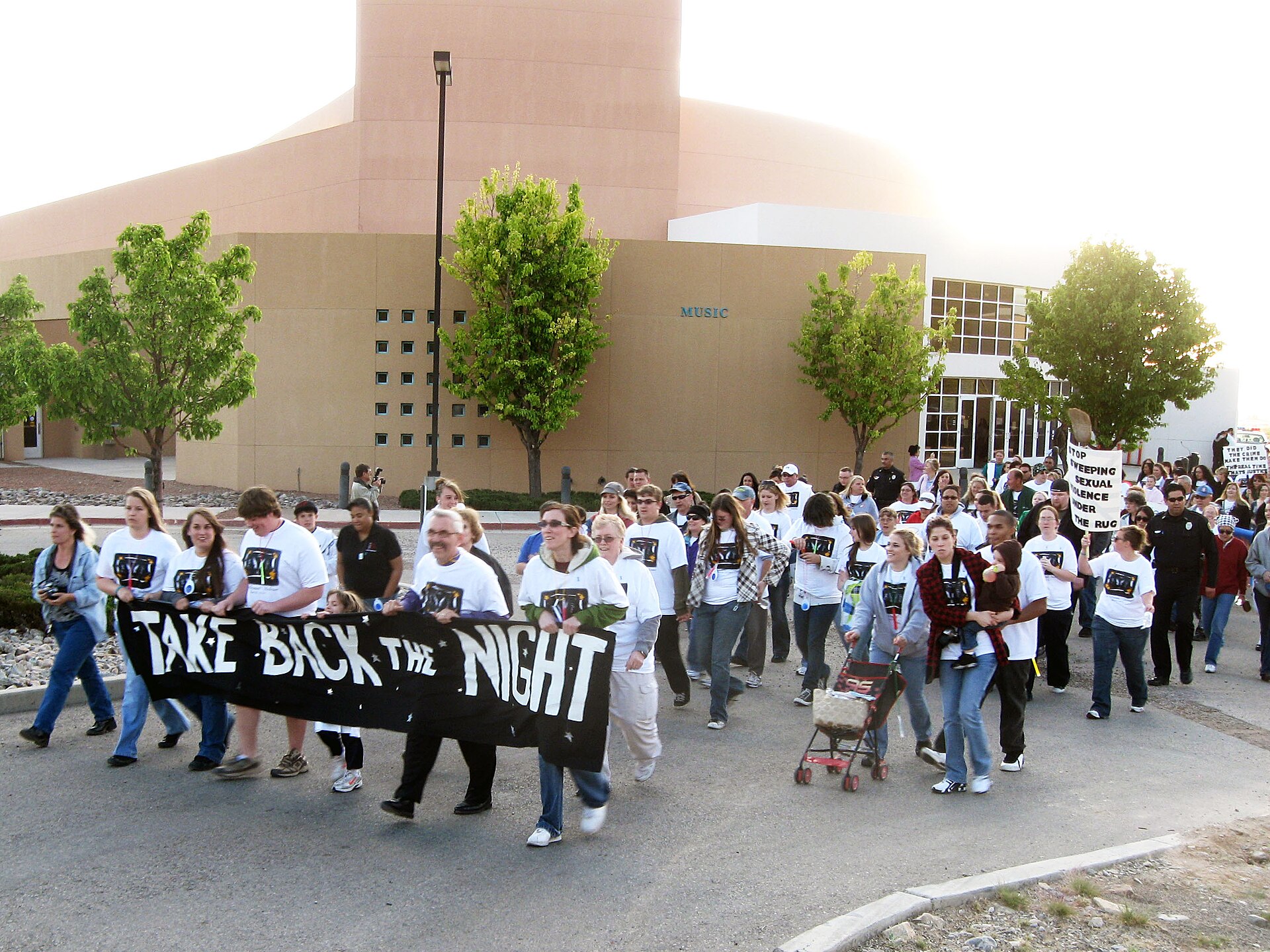 Fotografía en color de una marcha nocturna de centenares de personas con camisetas blancas detrás de una pancarta negra con el texto Take Back the Night